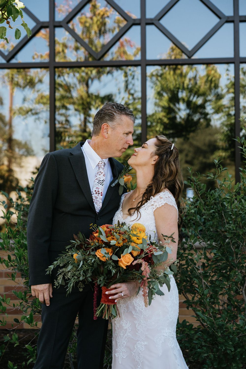 A Sacramento area bride and groom share a kiss in front of an ornate church window reflecting trees and a blue sky.