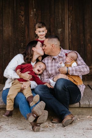 A husband and wife kiss while snuggling their three kids in front of a brown wooden barn door while posing for family photos.