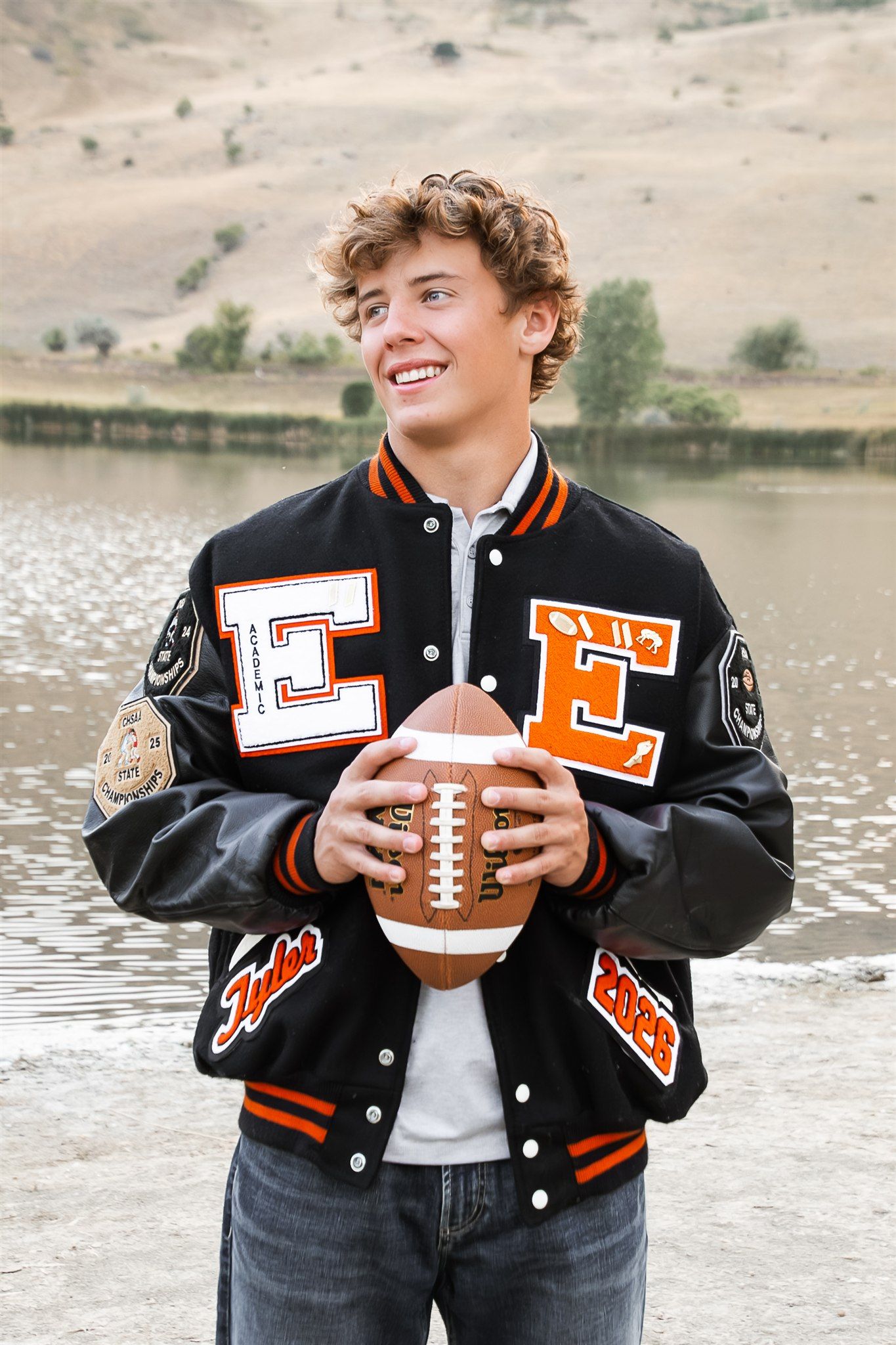 A football player in black uniform lies on the ground holding a football near rocky terrain.