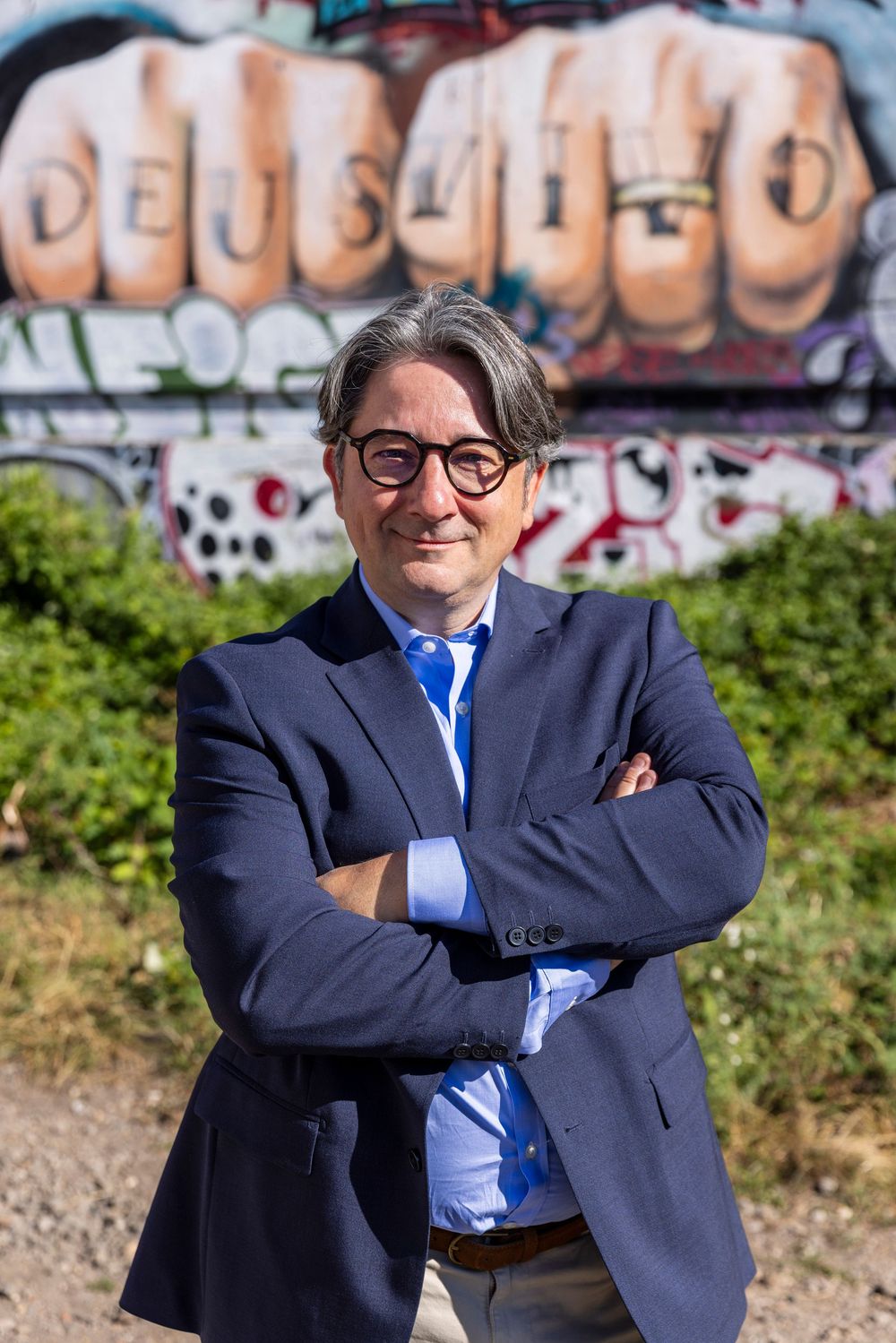 Professional business person in navy suit stands outdoors against graffiti wall background in casual poses.