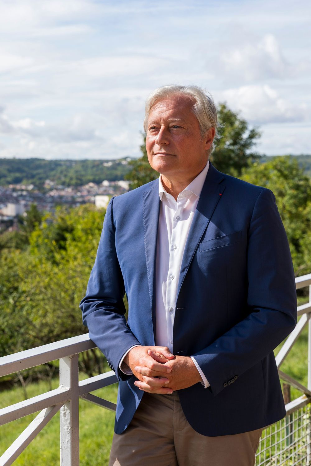 A person wearing a navy blazer and light pink shirt sits casually on a white railing with a scenic hilltop view.