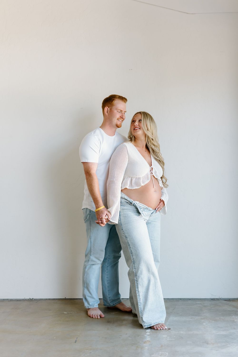 Couple in casual white and denim outfits standing together against white wall in maternity photo shoot.