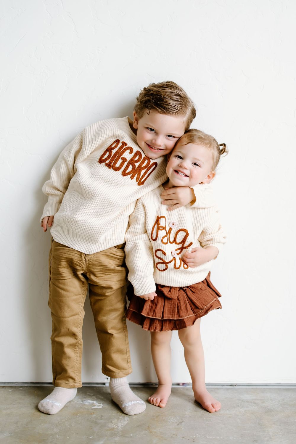 Two children wearing matching cream colored sweaters with text embrace against a white wall.