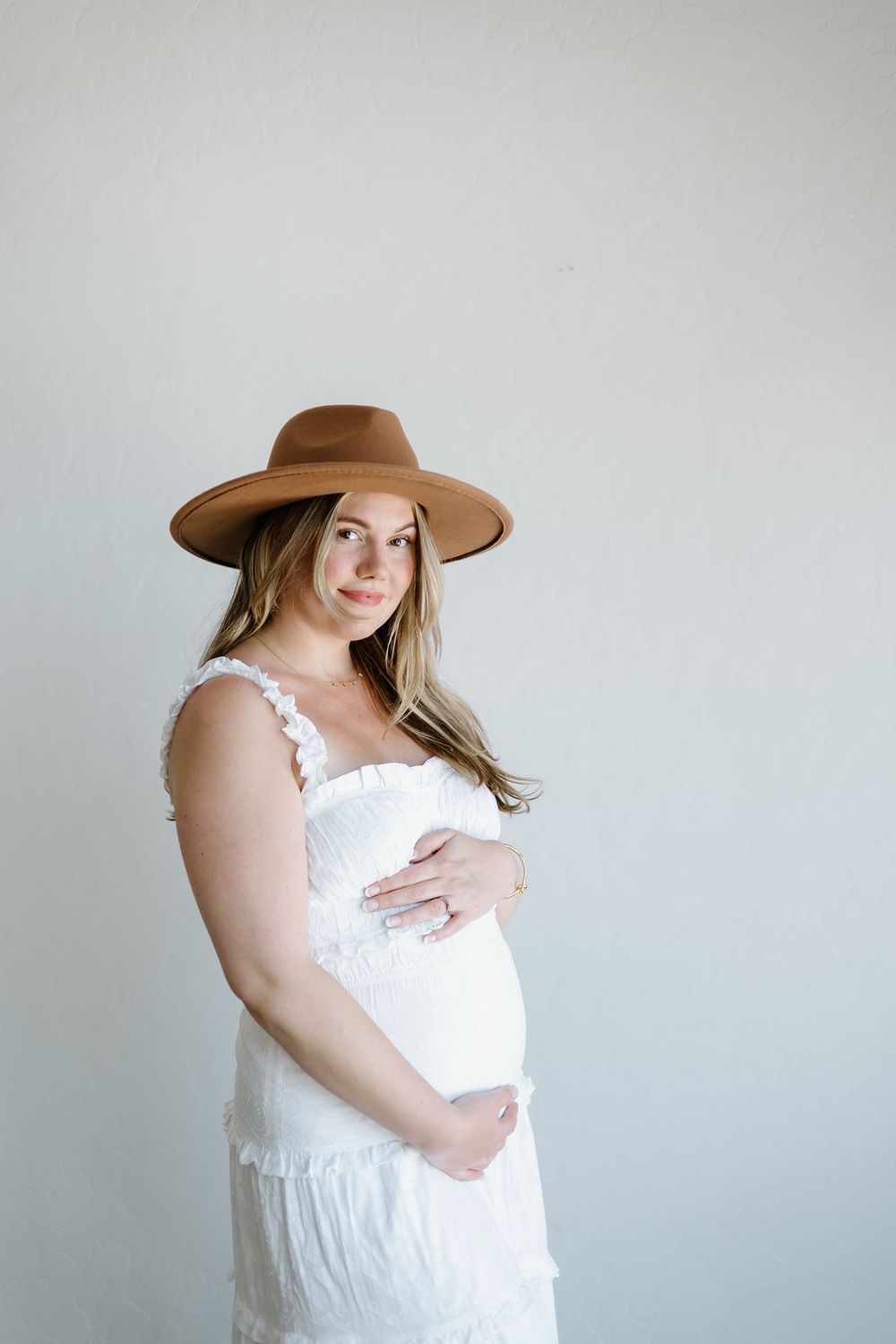 A woman in a brown hat and white dress poses against a light gray background.