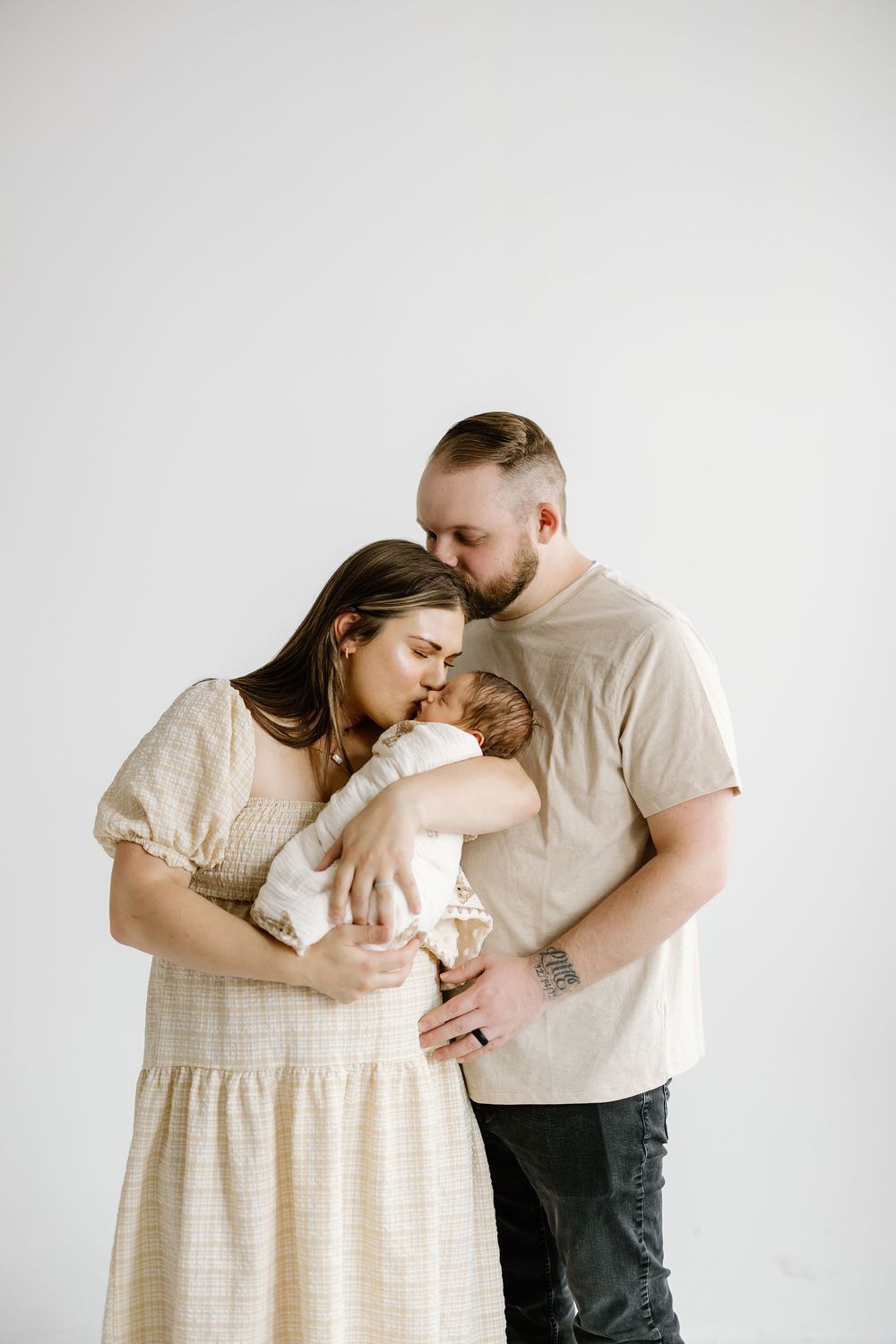 A couple in casual attire shares tender moments with their newborn baby against a white studio backdrop.