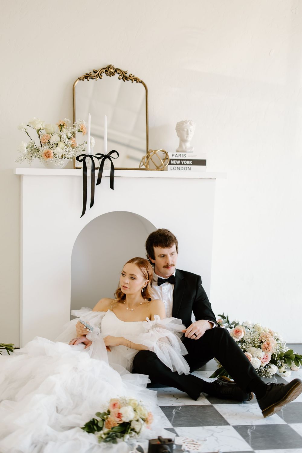 Couple in wedding attire relaxes on floor in bright white room with floral decor.