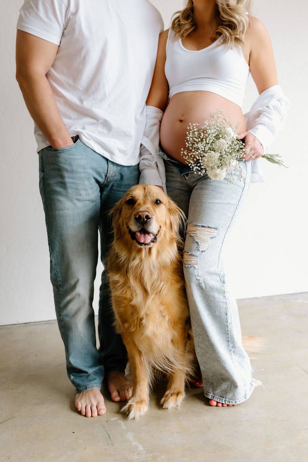 A couple in jeans and white shirts stands with their happy golden retriever against a white wall.