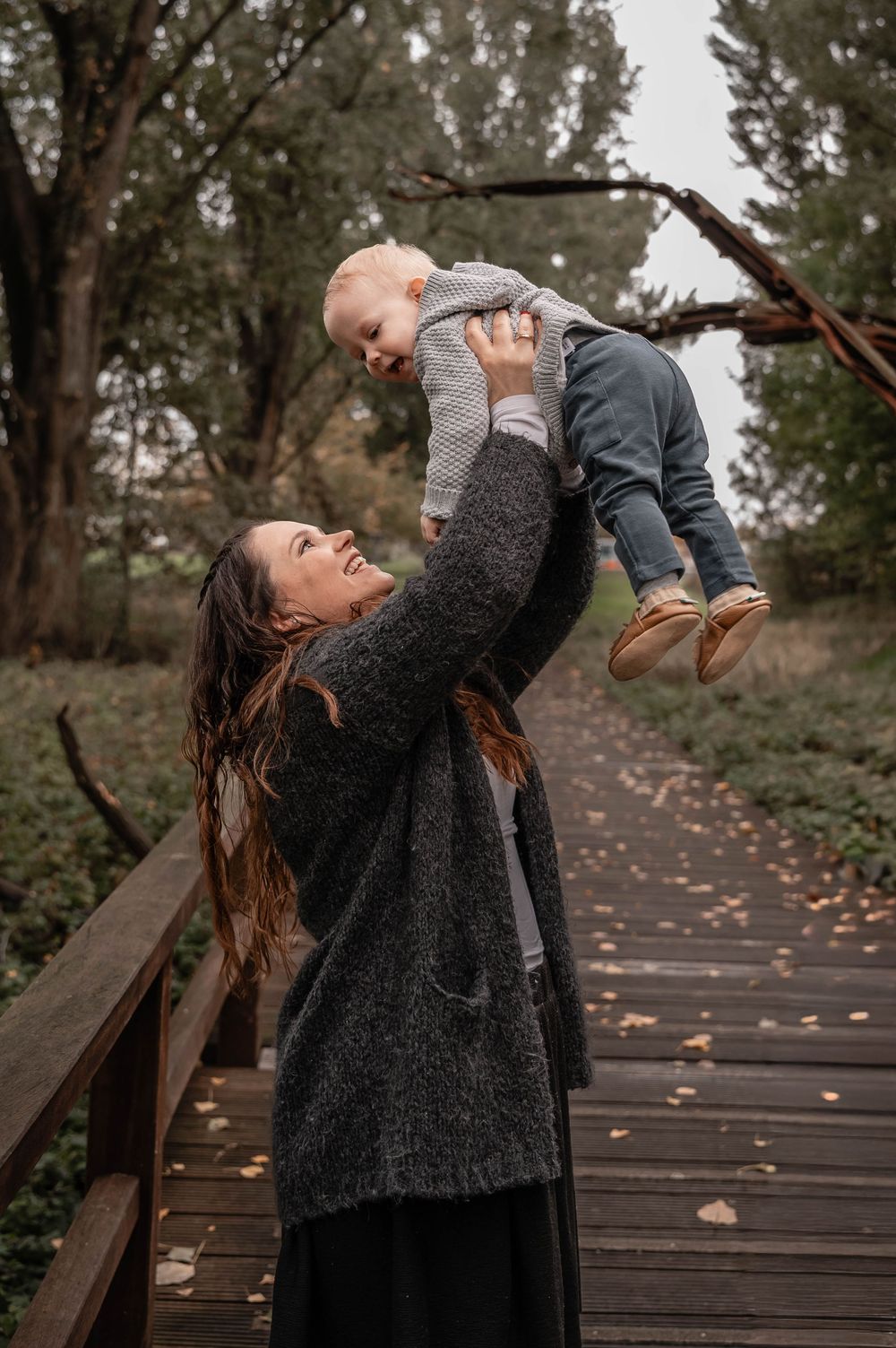 A joyful moment captured on a wooden boardwalk surrounded by autumn trees as a baby is lifted high into the air.
