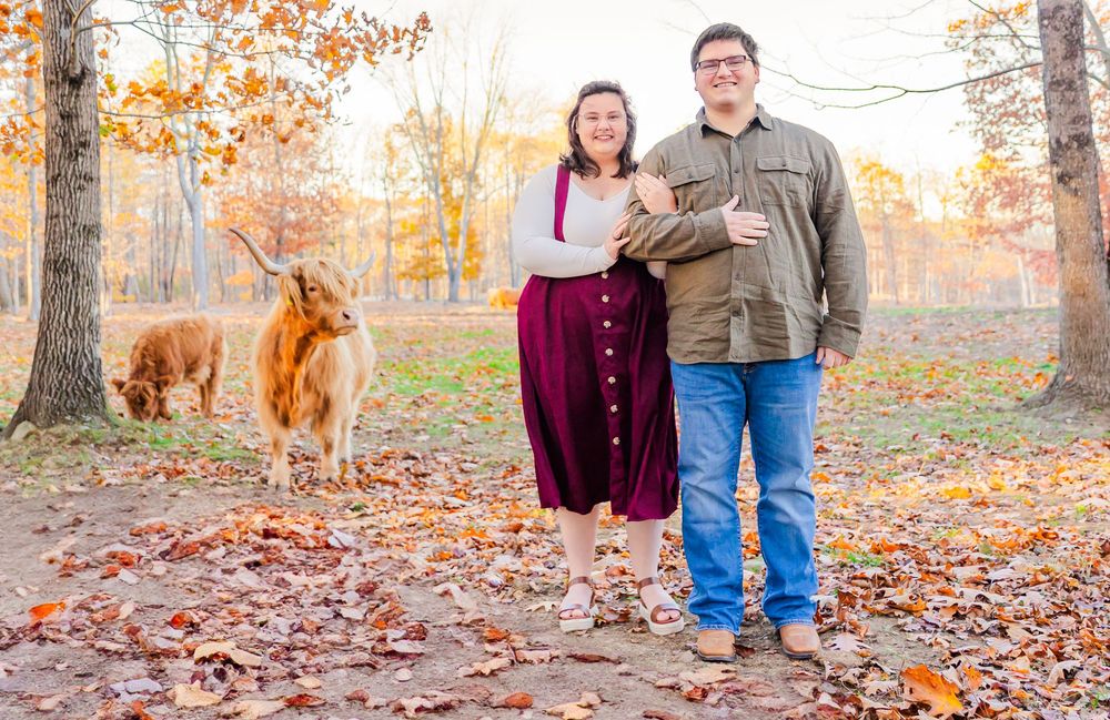 A couple and their golden retriever pose together on fallen autumn leaves in a scenic outdoor setting.