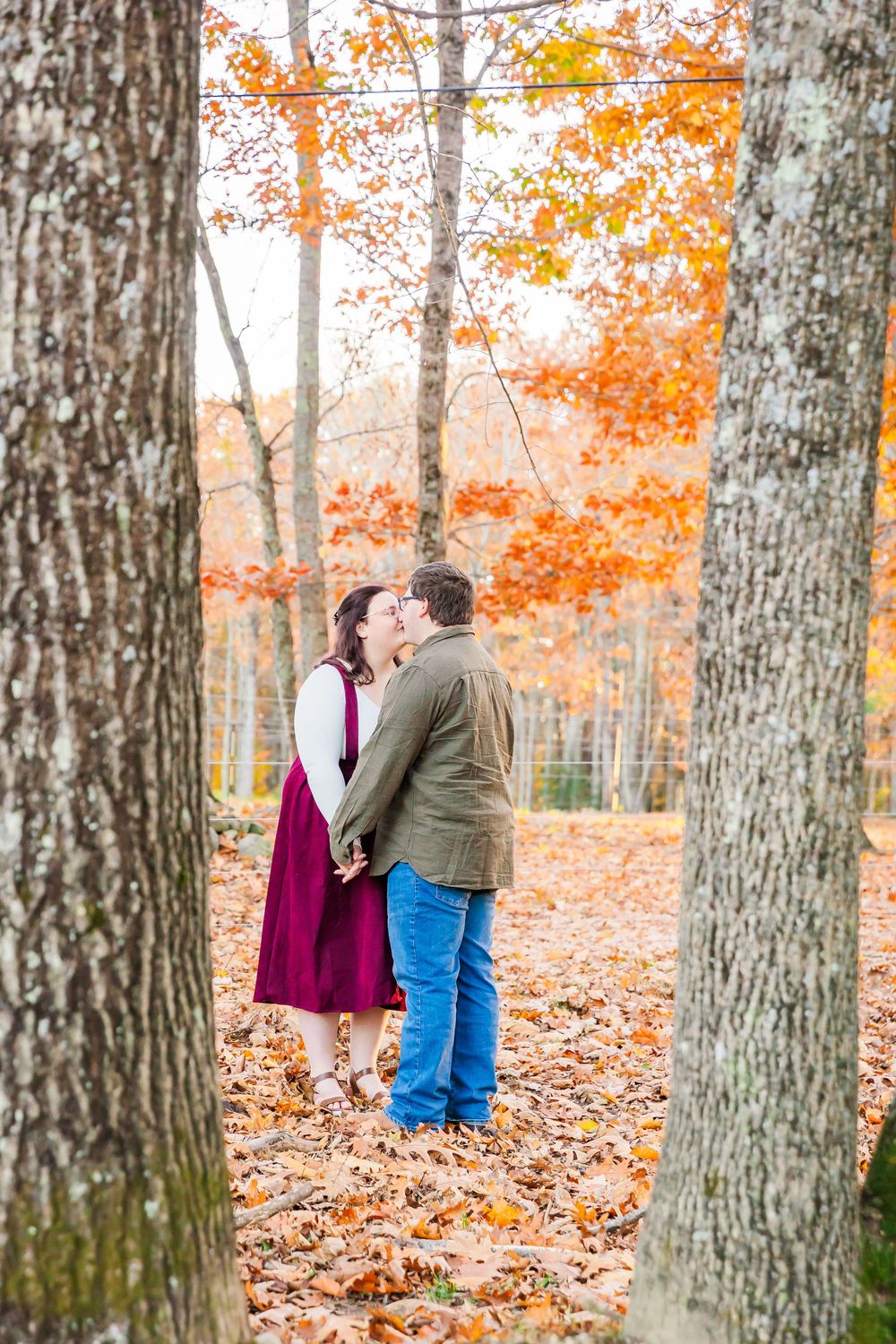 Two figures stand between tall trees in an autumn forest filled with orange and yellow leaves.