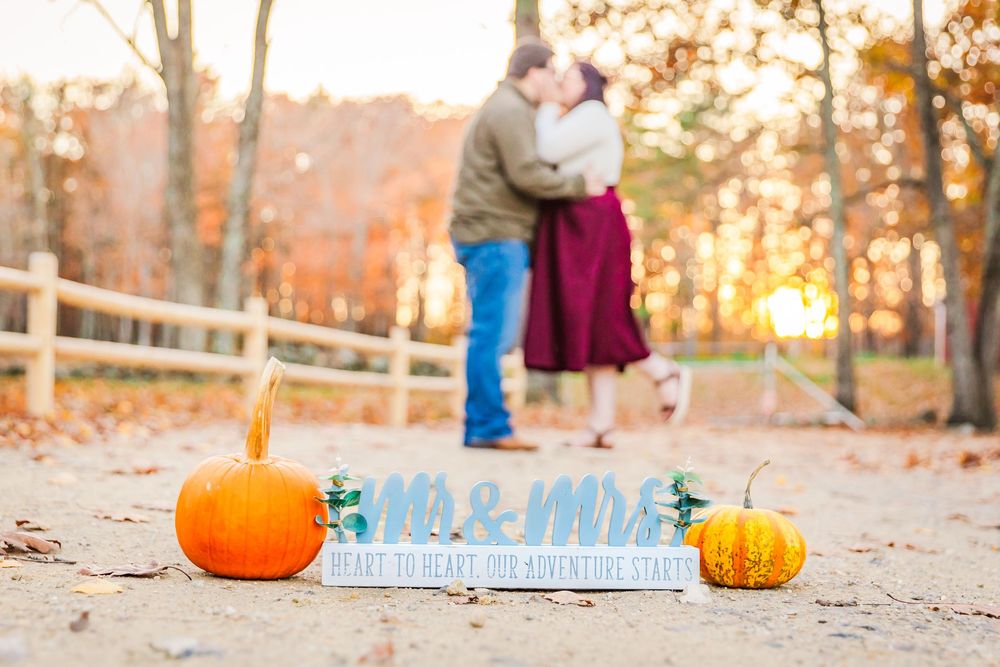A fall engagement announcement scene with two pumpkins and a blue 'Mr & Mrs' sign on rustic ground at sunset.