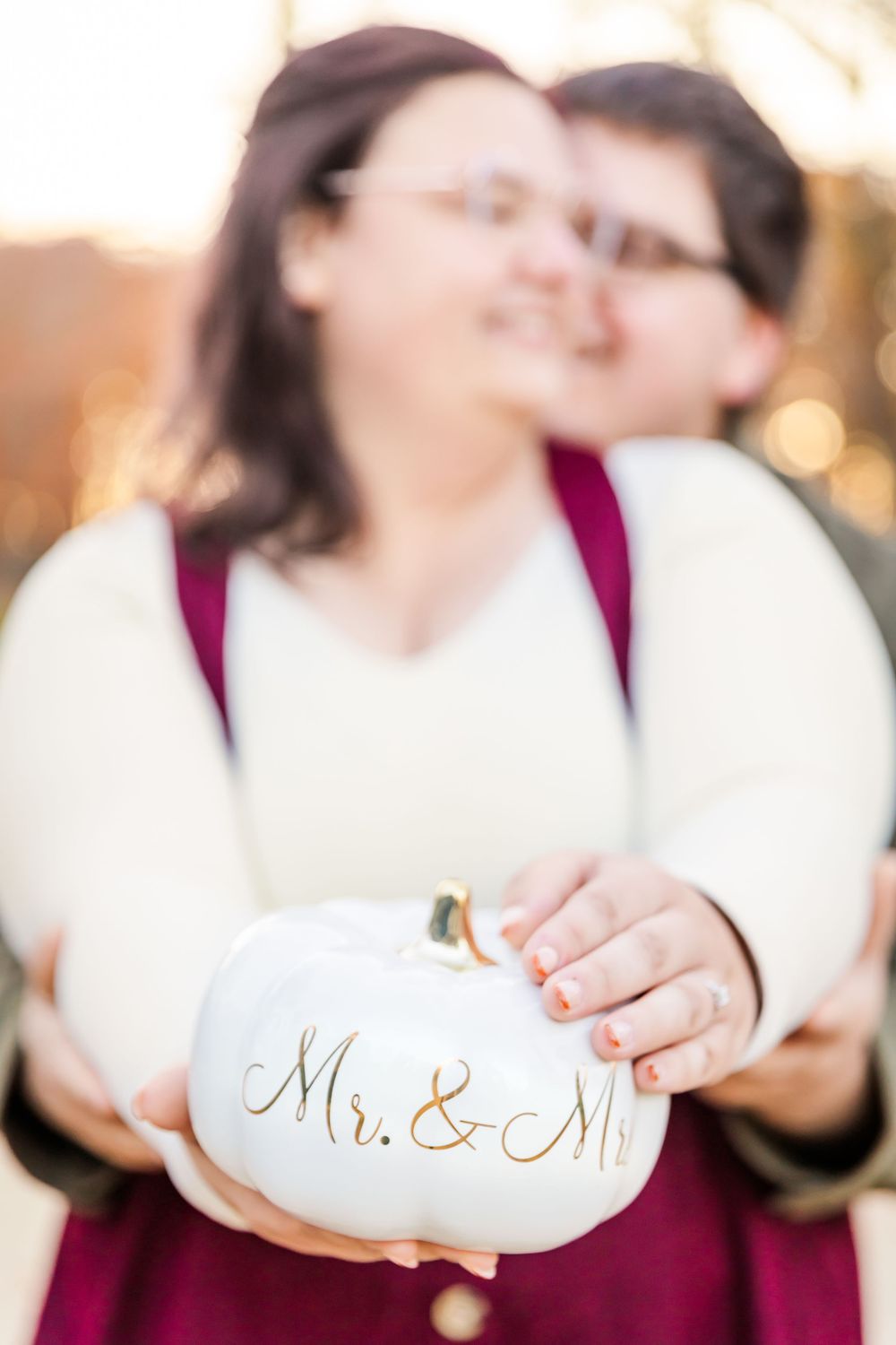 Hands hold a white decorative pumpkin with 'Mr & Mrs' text in an autumn outdoor setting.