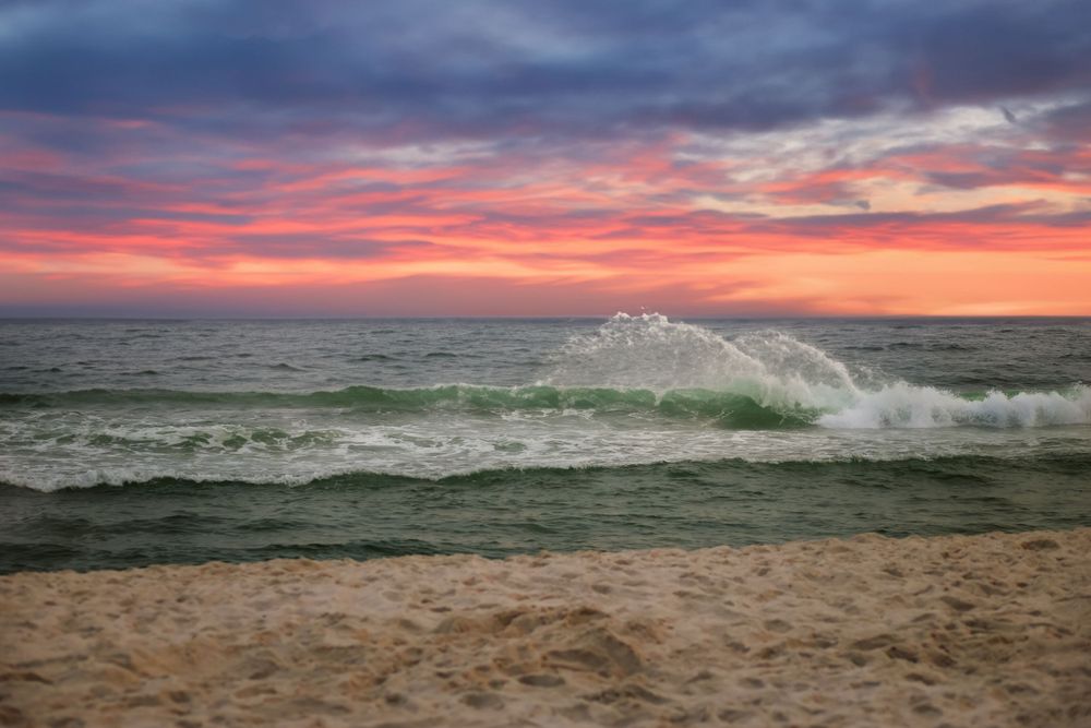 Vibrant sunset colors paint the sky as waves crash on sandy beach during golden hour.