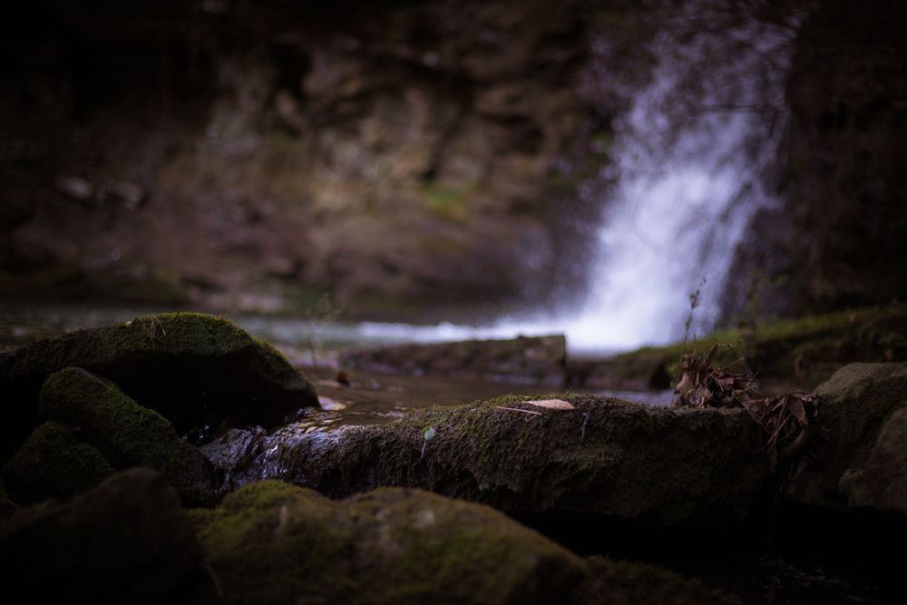 A misty waterfall splashes onto moss-covered rocks in dim lighting.