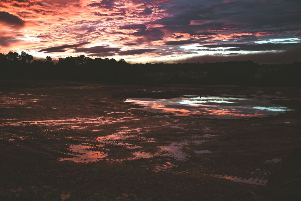 Dramatic red and orange sunset sky reflects off a still body of water with silhouetted treeline.