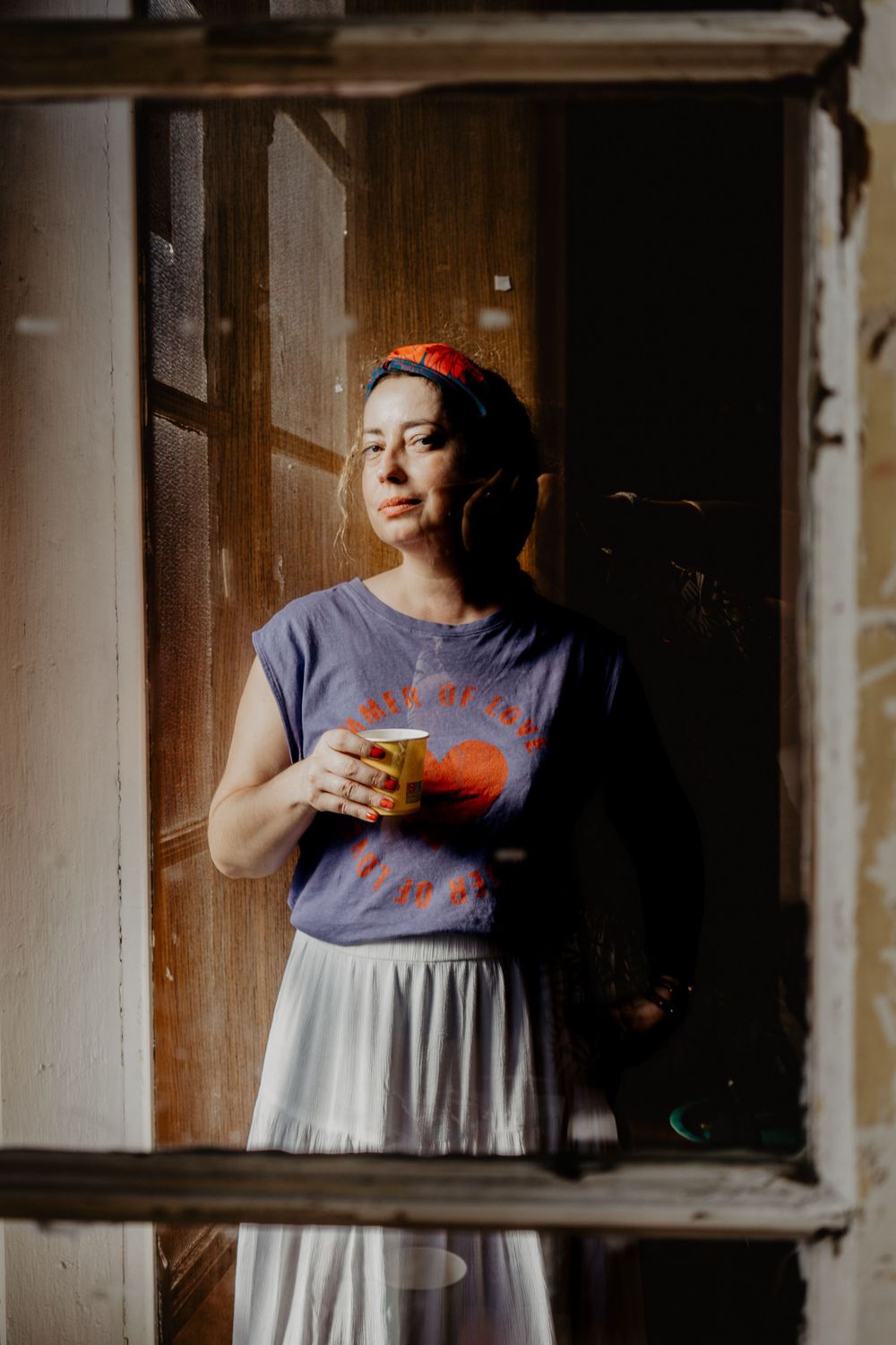 Retrato artístico de mulher em ambiente interior, observando pela janela com expressão contemplativa e segurando uma bebida