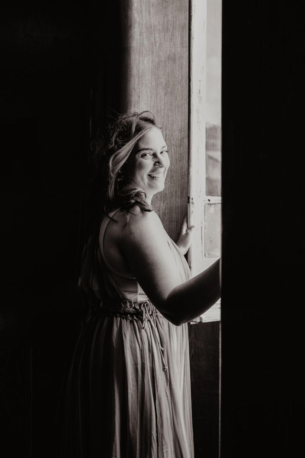 Retrato em preto e branco de mulher sorrindo suavemente enquanto olha pela janela, iluminada por luz natural suave.