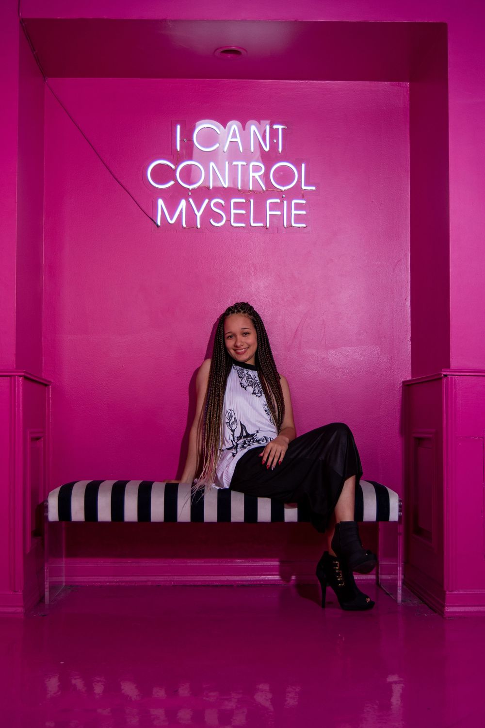 Pink neon sign reading I cant control myselfie above striped bench in pink room.