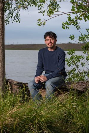 Young man in navy sweater and jeans sitting on a log by a lake under a tree in a green outdoor setting.