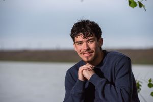 Young man with dark hair smiling outdoors near a lake, wearing a navy sweatshirt on a cloudy day.