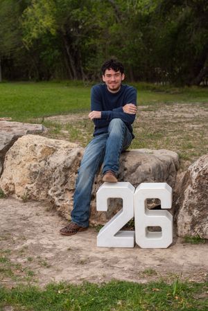 Young man sitting on rocks next to large white number 26 sign in a park setting.