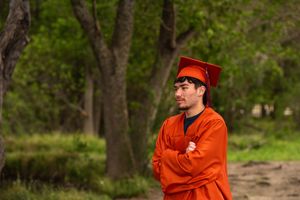 Young man in orange graduation cap and gown standing outdoors with arms crossed among green trees.