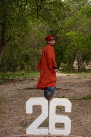Graduate in red cap and gown standing by large white number 26 outdoors.
