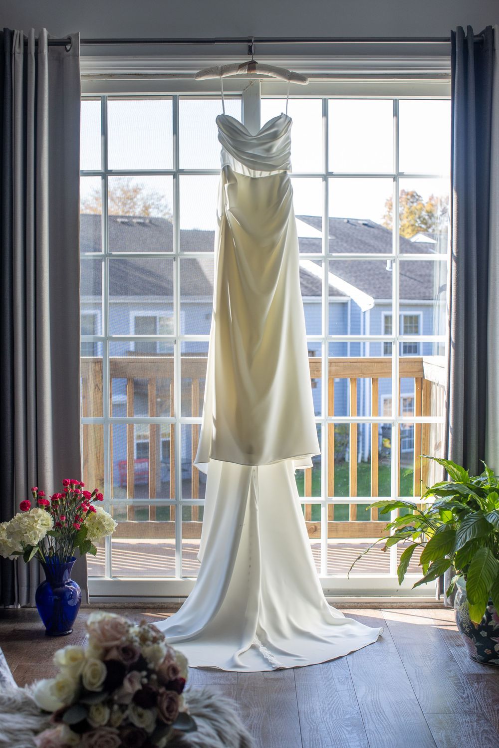 Elegant ivory satin wedding gown hanging in sunlit window with floral arrangements and bouquet below.