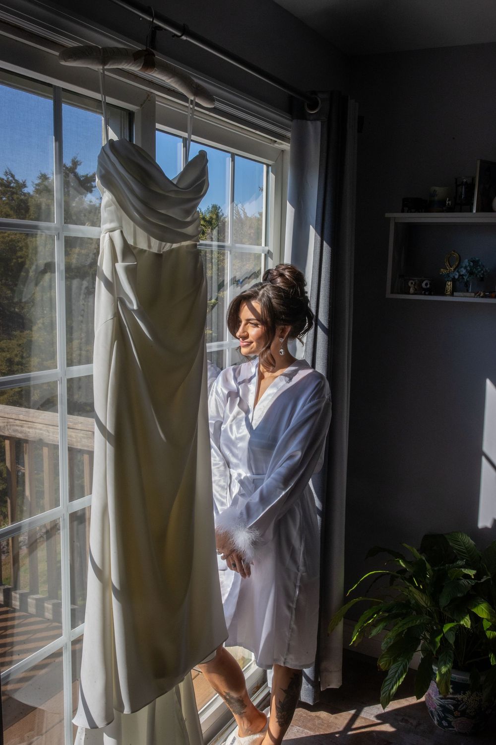 Bride in white robe standing beside her hanging wedding gown in a sunlit window before the ceremony.