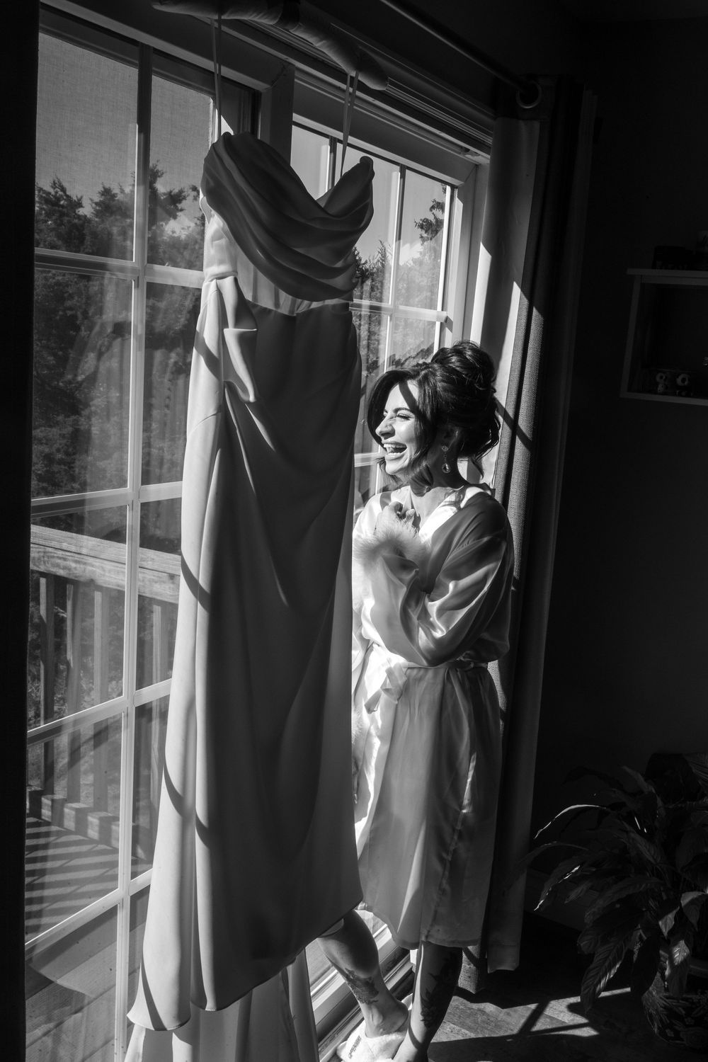 Black and white photo of laughing bride in robe standing beside her hanging wedding gown in window light.