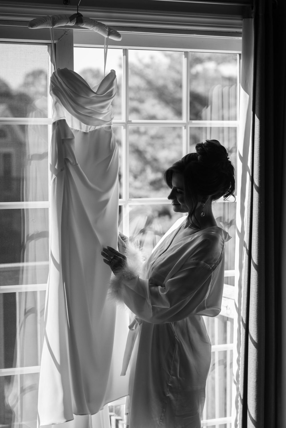 Elegant black and white silhouette of bride in robe admiring her wedding gown hanging in a sunlit window.