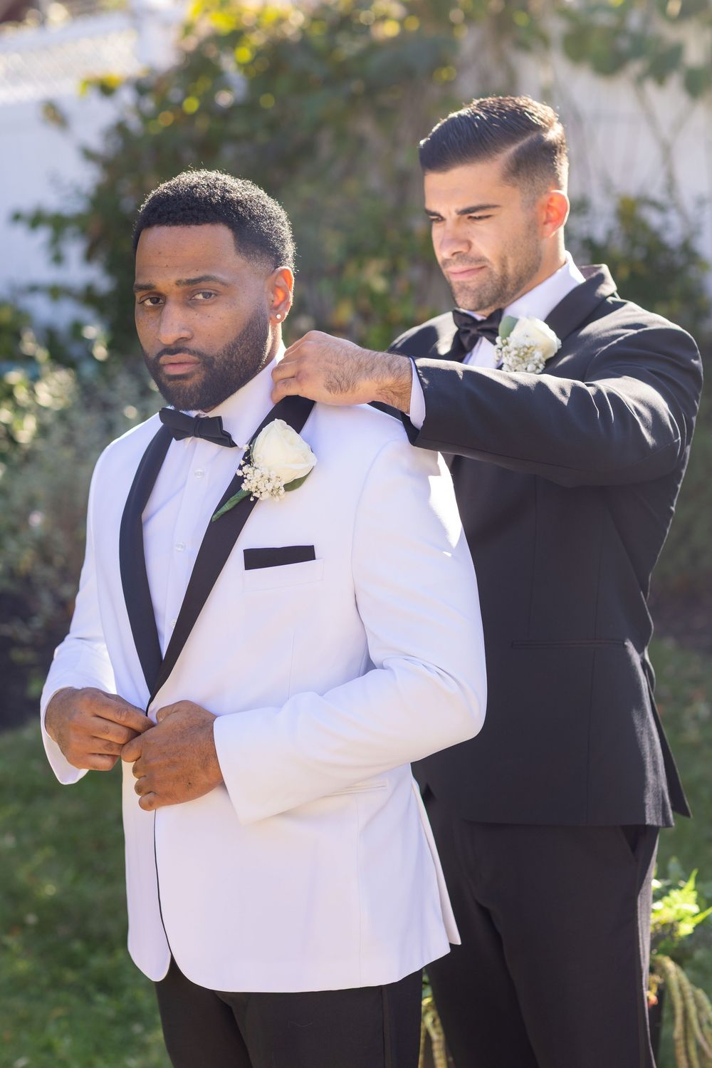 Groomsman adjusting the collar of groom in white tuxedo jacket outdoors.