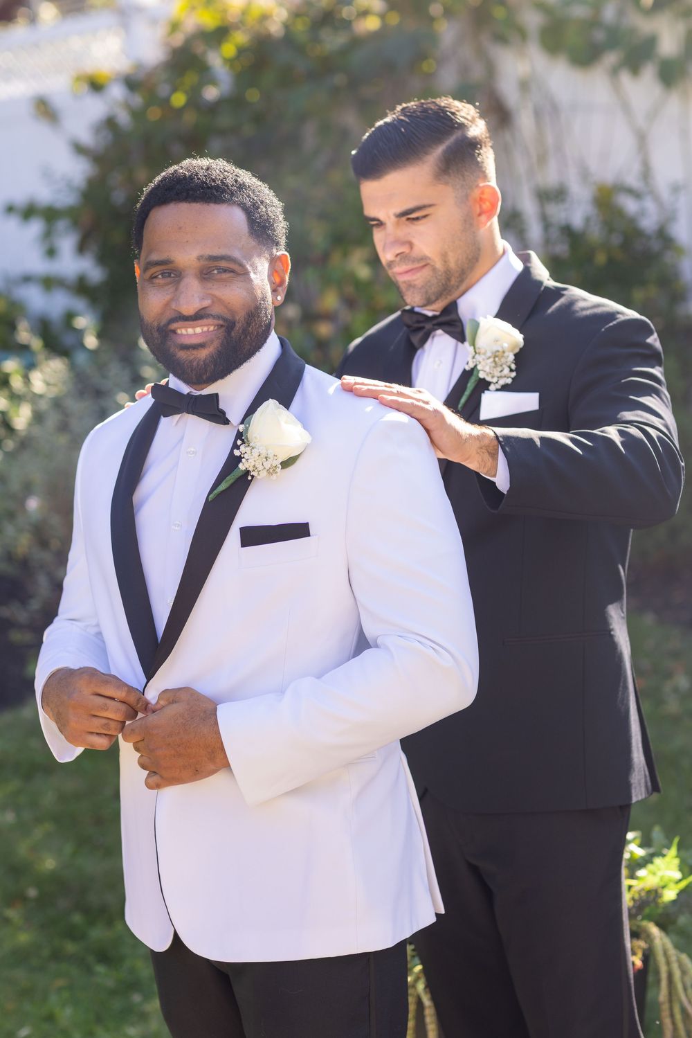 Smiling groom in white tuxedo being assisted by groomsman in black suit outdoors.