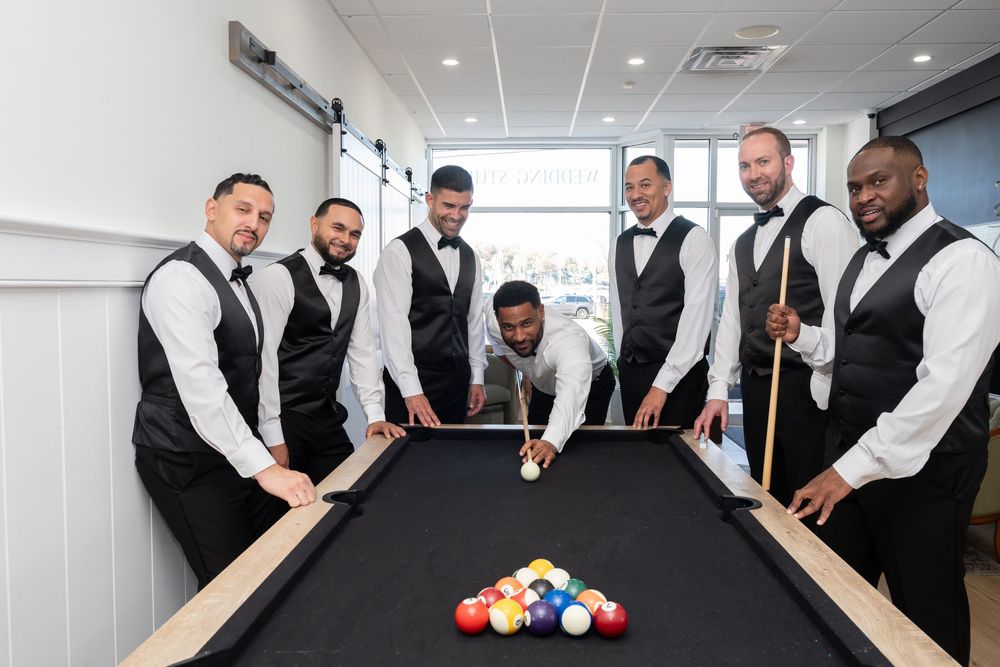 Groom and groomsmen in black vests gathered around a pool table for a fun wedding photo.