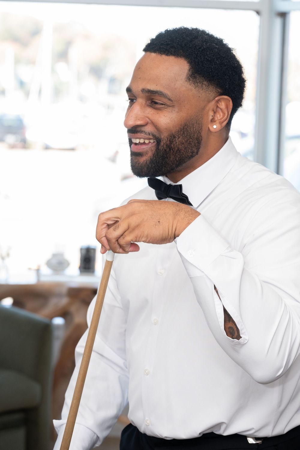 Smiling groom in white shirt and black bow tie leaning on a pool cue at wedding venue.