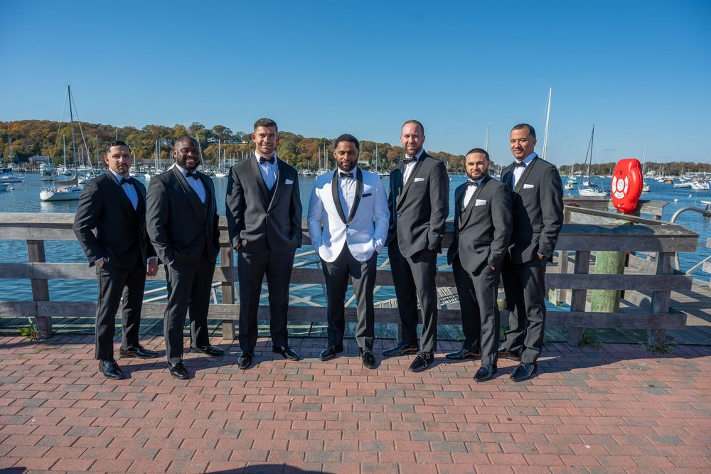 Groom in white tuxedo jacket with groomsmen in dark suits posing at waterfront marina.
