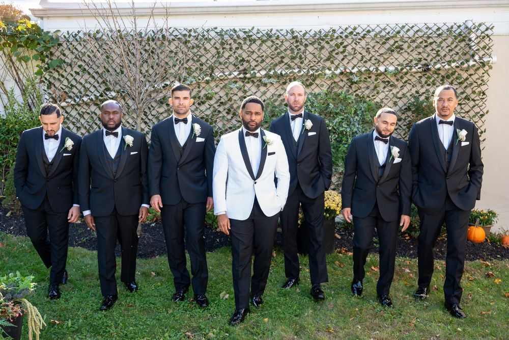 Groom in white tuxedo jacket stands with seven groomsmen in black tuxedos outdoors at a wedding.