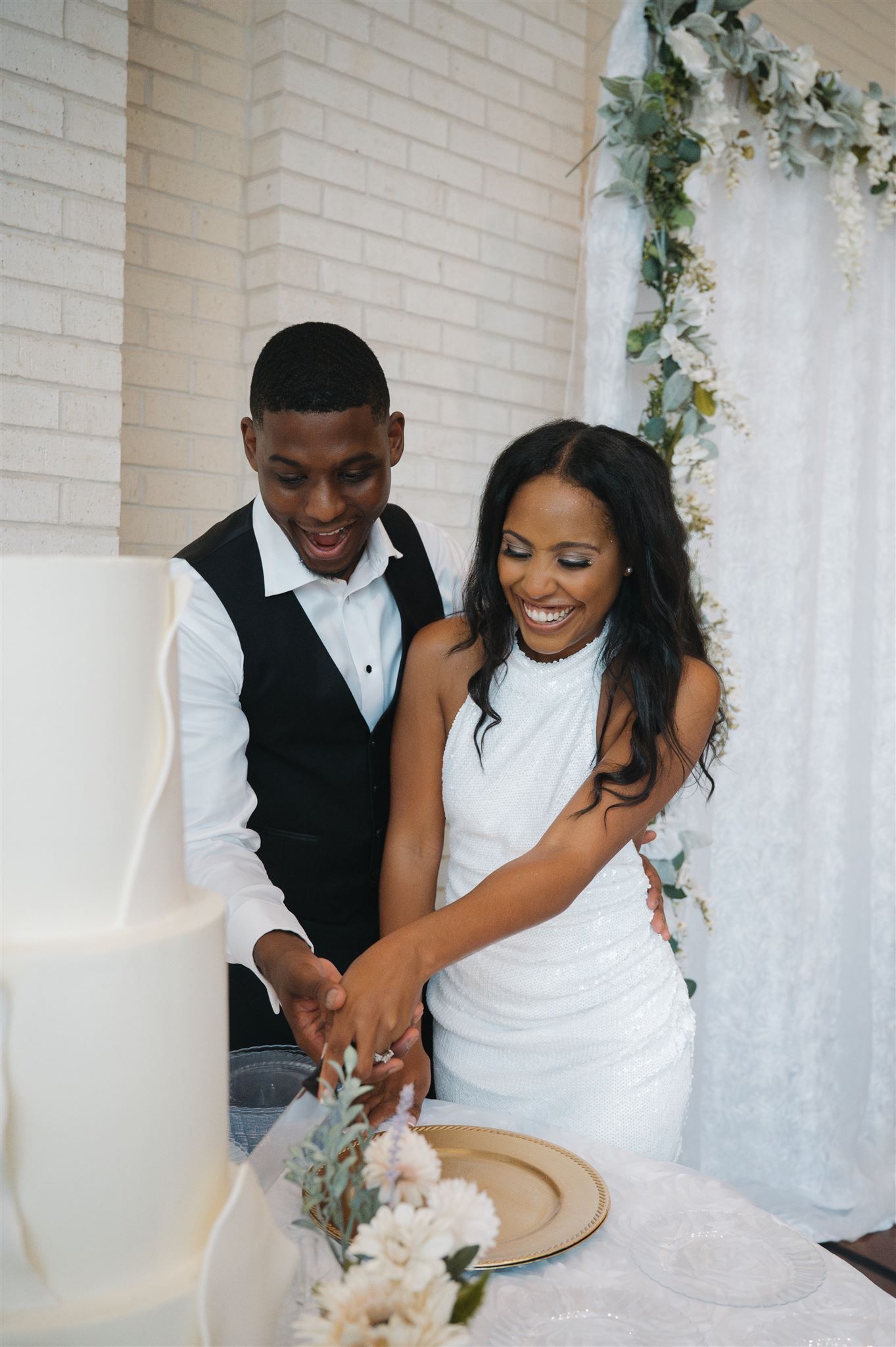 A happy couple in formal wedding attire laugh while cutting their elegant white wedding cake together.