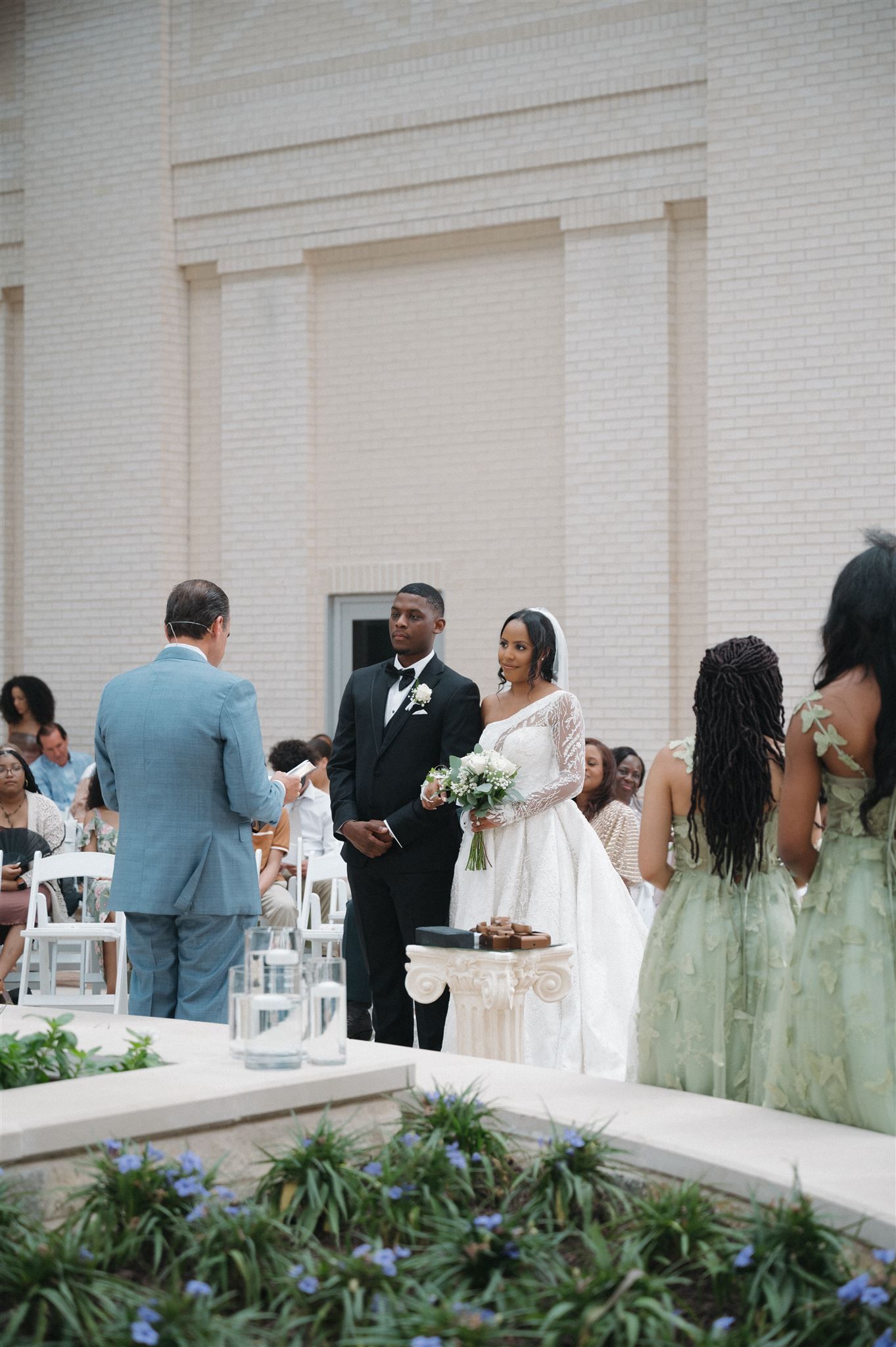 Wedding guests gather for an outdoor ceremony in front of a white brick building with mint green bridesmaids' dresses.