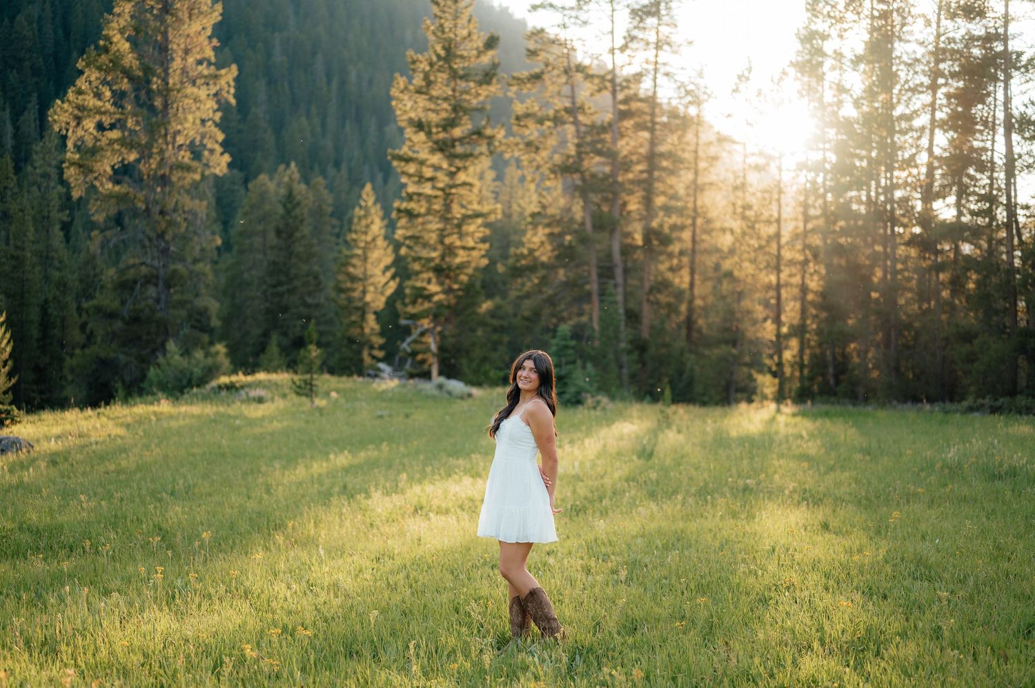 Young woman in white dress and cowboy boots standing in a sunlit mountain meadow at golden hour.