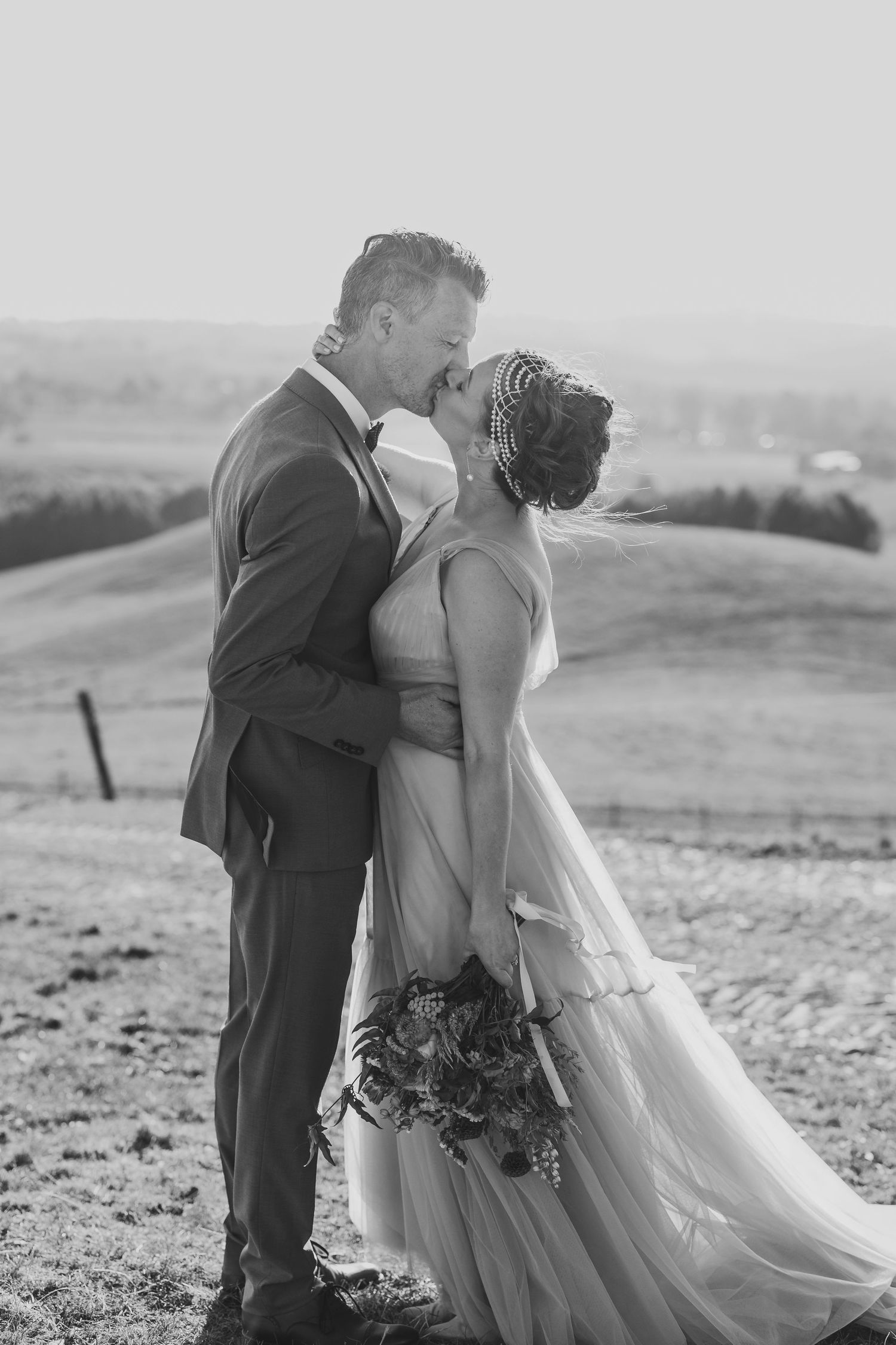 A couple shares an intimate moment in formal wedding attire on a hilltop with scenic countryside views.