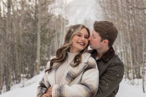 A couple shares an intimate moment in a snowy forest while wearing cozy winter sweaters.