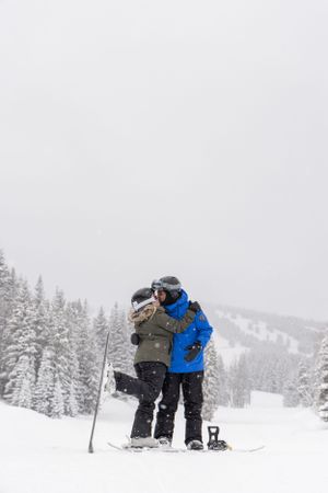 Two skiers embrace on a snowy mountain slope surrounded by evergreen trees in winter conditions.