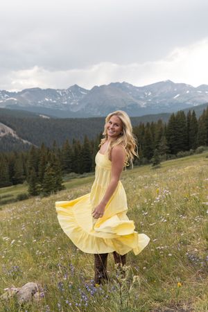 A yellow sundress twirls against a scenic mountain backdrop with wildflowers and evergreen trees in Colorado.