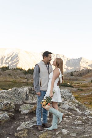 A couple stands together on rocky terrain with snow-capped mountains in the background at sunset.