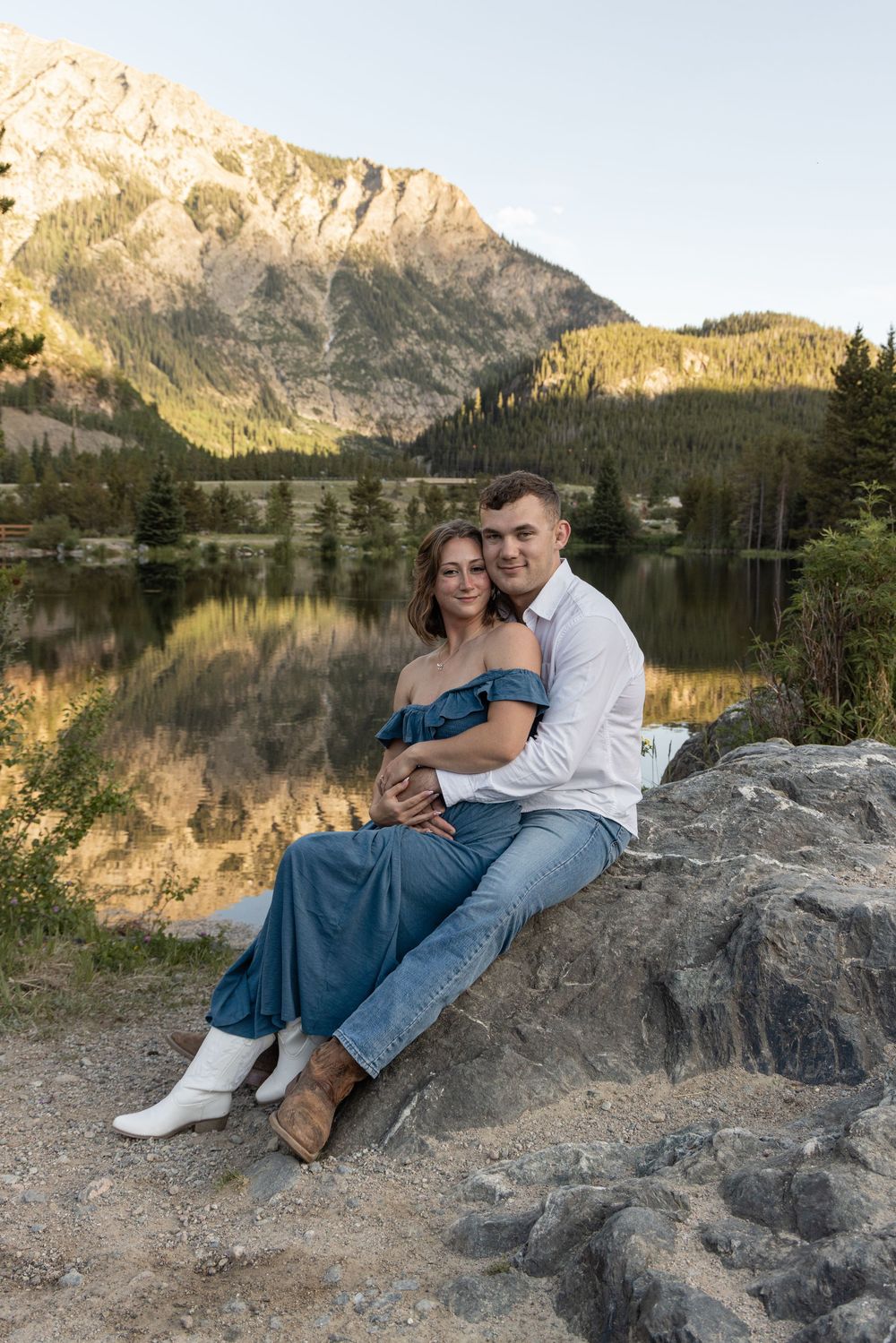 A couple sits on lakeside rocks at sunset with mountain reflections in Officer's Gulch