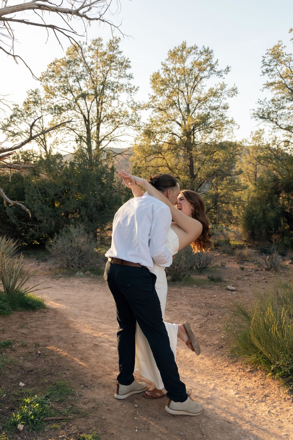 A romantic couple shares a tender embrace on a desert trail surrounded by desert brush and trees at sunset.