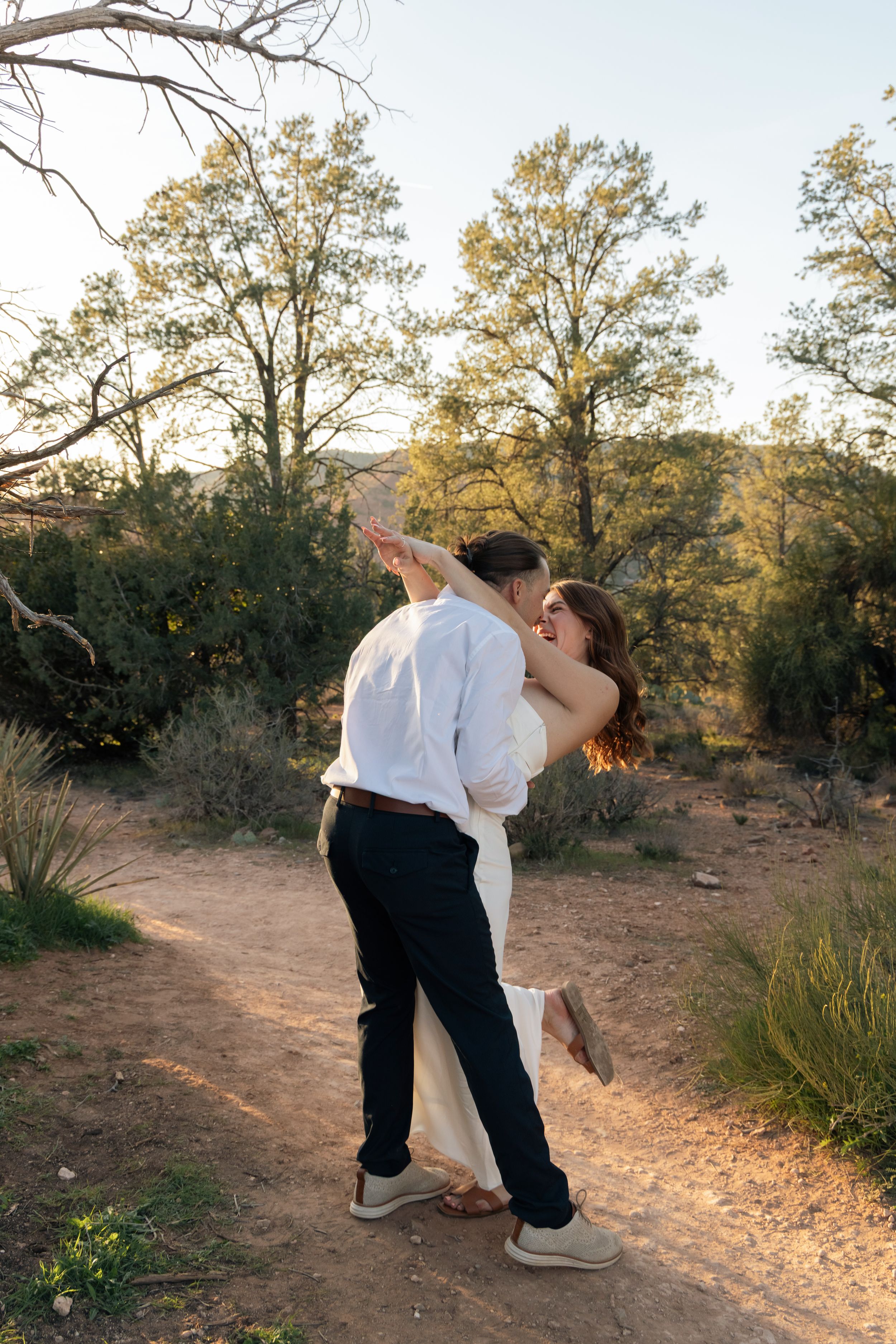 A romantic couple shares a tender embrace on a desert trail surrounded by desert brush and trees at sunset.