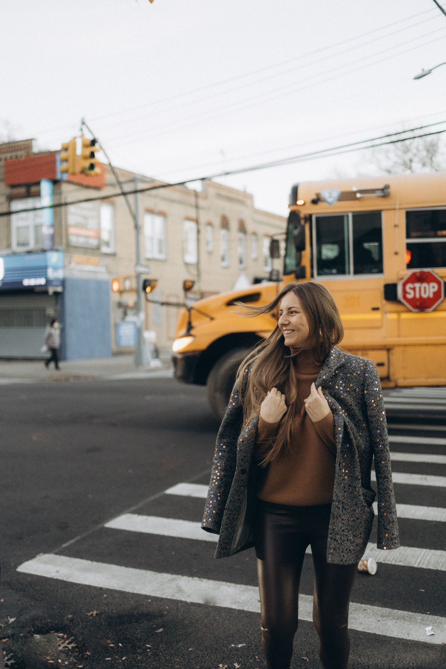 Smiling woman in gray coat crossing street with yellow school bus in urban background.