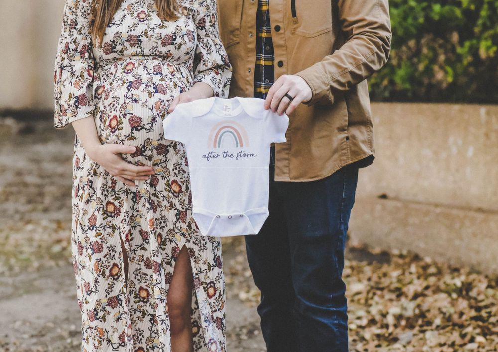 Expecting couple holding a rainbow baby onesie outdoors, woman in floral dress cradling her pregnant belly.